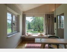 Innenraum eines modernen Wohnzimmers mit hellen Holzwänden und -decken. Im Mittelpunkt steht eine große Fensterfront mit Blick auf einen grünen Garten, davor eine Fensterbank mit Sitzkissen. Ein Holztisch mit einer Blumenvase und Teppich ergänzen das skandinavisch inspirierte Design.