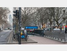 U-Bahneingang zur Haltestelle "Uhlandstraße" in Berlin, umgeben von einer belebten Straße mit geparkten Fahrrädern, Geschäften und Bäumen. Im Hintergrund ist die Kaiser-Wilhelm-Gedächtniskirche sichtbar.