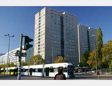 Mehrstöckiger Wohnblock aus Betonplatten in urbaner Umgebung, davor eine Straßenbahn mit Werbung, Fußgänger und eine Ampel. Im Hintergrund sind Bäume und weitere Gebäude unter klarem, blauem Himmel zu sehen.