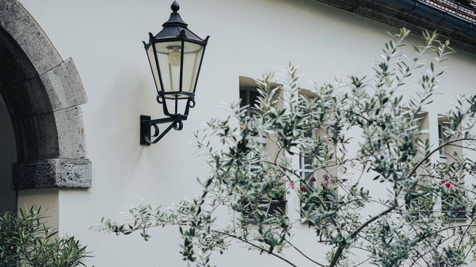 Außenansicht eines hell verputzten Gebäudes mit schwarzer, klassischer Wandlaterne und einem steinernen Rundbogen am linken Rand. Im Vordergrund stehen Zweige eines Baumes; dahinter sind Fenster mit Blumenkästen unter einem Dach mit Regenrinne zu sehen.