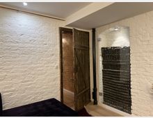 Raum mit weiß gestrichenen Ziegelwänden, einer Holztür im rustikalen Stil und einem großen Wandspiegel, der schwarze gestapelte Kacheln reflektiert. Der Boden ist aus hellem Holz, und eine schwarze Sitzfläche ist teilweise sichtbar im Vordergrund.