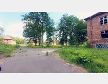 Eine verlassene Anlage mit mehreren backsteinernen Gebäuden, umgeben von hohen Bäumen und überwucherndem Gras. Der Boden besteht aus Rissen durchzogenen Asphaltflächen, und eines der Gebäude zeigt Graffiti an der Fassade.
