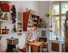 Eine kleine Küche mit hellen Wänden, offenen Regalen und einer Hängelampe. Links stehen Küchenutensilien und Gewürze auf Regalen, zentral ein Holzwandregal mit Geschirr und Büchern, und rechts ein Tisch mit Stuhl neben einem großen Fenster, durch das ein Hof mit Bäumen sichtbar ist.