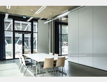 Moderner Besprechungsraum mit einem weißen Tisch und mehreren Holzstühlen, umgeben von großen Glasfenstern mit Blick nach draußen. Die Wände sind schlicht und hell, die Deckenleuchten ergänzen das minimalistische Design.