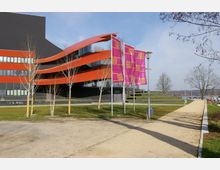 Modernes Gebäude mit schwarz-roter Fassade und geschwungenen roten Elementen, umgeben von einer Freifläche mit jung gepflanzten Bäumen und einem Gehweg. Im Vordergrund wehen pinke Fahnen mit gelber Aufschrift "Schiffbauergasse", und im Hintergrund sind Rasenstufen sowie eine Flusslandschaft zu erkennen.