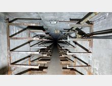 Ein enger, unterirdischer Betonraum mit rostigen Metallträgern, die von den Wänden ragen, und einer Lampe an der Decke. Der Raum wirkt ungenutzt und zeigt Abnutzungserscheinungen.