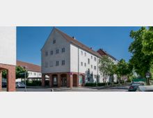 Mehrgeschossiges Wohngebäude im traditionellen Stil mit weiß verputzten Wänden, rotem Satteldach und einem Erdgeschoss mit Backsteinfassade. Die Umgebung zeigt eine ruhige Straße mit Autos, gepflastertem Bürgersteig und Bäumen.