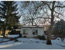 Schneebedeckter Garten mit einem kleinen weißen Gebäude im Hintergrund, umgeben von kahlen Bäumen und immergrünen Nadelbäumen. Im Vordergrund ist ein verschneiter Weg sichtbar, sowie ein großer Baum mit einer Schaukel.