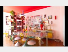 Wohnzimmer mit einer auffälligen, bunten Einrichtung. Ein verspiegelter Esstisch im Vordergrund, umgeben von Stühlen mit gebogenen Metalllehnen, eine rosa Regalwand mit Büchern und Pflanzen auf der linken Seite sowie eine helle Wand mit moderner Kunst und Dekorationen im Hintergrund.