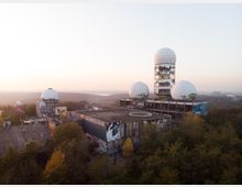 Ehemalige Abhörstation auf einem Hügel, bestehend aus mehreren runden Kuppeln mit Wabenmuster, die auf teils zerfallenen und mit Graffiti bedeckten Gebäuden stehen, umgeben von bewaldeter Landschaft in der Abenddämmerung.