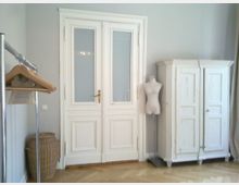 Heller Raum mit einer Doppeltür aus weiß gestrichenem Holz und mattem Glaseinsatz in der Mitte. Auf der rechten Seite ein weißer Vintage-Schrank, daneben eine Schneiderpuppe; links ein Korb und eine Kleiderstange mit Holzbügeln.