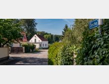 Eine Dorfstraße mit einem unbefestigten Weg, gesäumt von dichten Hecken und Bäumen. Im Hintergrund sind weiße Häuser mit roten Ziegeldächern, umgeben von grüner Vegetation, zu sehen; ein Straßenschild mit der Aufschrift „Deiche“ steht im Vordergrund.