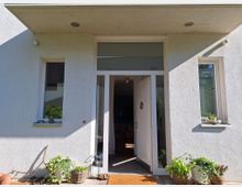 Moderne Hauseingangsfassade mit weißer Tür und großen Glasfenstern. Auf beiden Seiten befinden sich rechteckige Fenster, daneben verschiedene Pflanzen in Töpfen sowie dekorative Gegenstände.