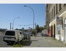 Straßenszene mit breitem Gehweg, geparkten Fahrzeugen und einem mehrstöckigen Gebäude mit gelblicher Fassade und bogenförmigen Eingängen. Im Hintergrund sind Straßenlaternen, Oberleitungen und vereinzelt moderne Gebäude sichtbar, während rechts am Gehweg Menschen an Tischen sitzen.