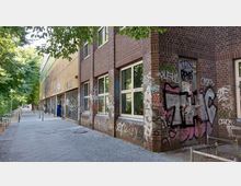 Backsteinbau entlang eines Gehwegs in einer städtischen Umgebung, mit großen Fenstern und Wänden, die mit Graffiti bedeckt sind; im Hintergrund sind Bäume zu sehen, die Schatten spenden, sowie Fahrradständer und Straßenlaternen.