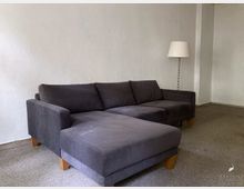 Graues Sofa mit Holzfüßen in einem schlicht eingerichteten Raum mit weißer Wand und Teppichboden; im Hintergrund steht eine Stehlampe mit weißem Lampenschirm.