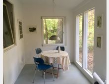 Kleiner, heller Raum mit großen Fenstern, die auf einen bewaldeten Garten blicken. Ein runder Tisch mit gemusterter Tischdecke, drei blauen Stühlen und einer Pendelleuchte darüber befindet sich in der Mitte; an den Wänden hängen minimalistische Bilder.