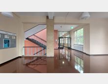 Flur eines Schulgebäudes mit glänzendem braunem Boden, großen Fenstern auf der rechten Seite, einer Glasdoppeltür im Hintergrund und einer Treppe mit Metallgeländer auf der linken Seite. Die Wände sind beige, mit einer Pinnwand links und einem roten Treppenhausbereich.