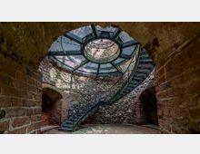 Innenraum eines historischen, runden Turms mit unverputztem Mauerwerk aus Stein und Backstein. Eine Metallwendeltreppe führt zu einer verglasten Kuppel, die Licht hereinlässt.