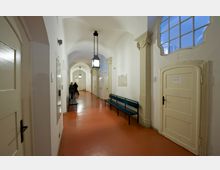 Ein Flur mit gewölbter Decke in einem historischen Gebäude, ausgestattet mit roten Böden, weißen Wänden, hölzernen Türen und Sitzbänken entlang der Wände. Große Fenster und dekorative Säulen ergänzen die Architektur, während im Hintergrund Personen erkennbar sind, die sich unterhalten.