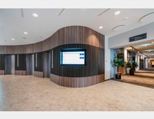 Moderner Innenbereich eines Gebäudes mit hellen Wänden, geschwungener Holzverkleidung und drei Aufzügen. Ein beleuchteter Bildschirm zur Orientierung ist zentral eingebaut, und ein Flur mit Teppichboden und Zimmerpflanzen führt weiter in den Hintergrund.