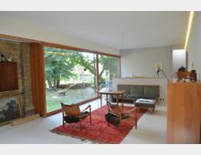 Wohnzimmer mit modernem Design, großen bodentiefen Fenstern, die den Blick auf einen grünen Garten freigeben. Der Raum ist mit Holzmöbeln, einem roten Teppich und einem gemauerten Kamin ausgestattet, wobei viel Tageslicht die helle und minimalistische Einrichtung hervorhebt.