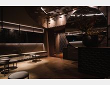 Ein modernes Innenraumdesign mit gedämpfter Beleuchtung, runden Tischen, wellenförmigen Wandverkleidungen und einem großen, runden Spiegel, der den Raum reflektiert. Eine Vase mit Zweigen und dezenten Pflanzen ergänzt das minimalistische Ambiente.