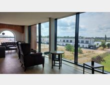 Gemütliche Lounge mit dunklen Ledersesseln und Tischen in einem Raum mit großen bodentiefen Fenstern, die den Blick auf moderne weiße Gebäude, eine weitläufige Grünfläche und einen Fluss freigeben. Links ein gemauerter Rundbogen mit Fenster im industriellen Stil.