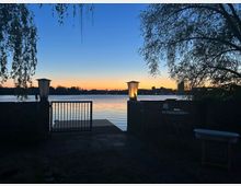 Blick von einer kleinen Uferterrasse mit Geländer und zwei Laternen auf einen ruhigen See bei Sonnenuntergang. Im Vordergrund rahmen Bäume und eine niedrige Mauer den Zugang zu einem kurzen Steg; am Horizont sind Silhouetten von Gebäuden zu sehen.