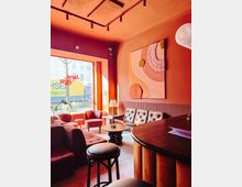Cozy café lounge with warm pink and orange walls, a large street-facing window, and a seating area with upholstered sofas and armchairs around small round tables. Track lighting hangs from the ceiling, and an abstract wall artwork is mounted above a tufted bench seat next to a curved wooden bar counter.