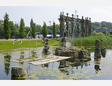 Ein Teich mit Seerosen, umgeben von skulpturalen Elementen aus Metall und Stein, sowie einer Ruinenarchitektur im Hintergrund. Im Umfeld befinden sich Grünflächen, Bäume und eine belebte Promenade.