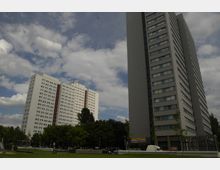 Zwei moderne Hochhäuser mit rechteckigen Fenstern, eines weiß mit roten Akzenten und eines grau, stehen neben einem bewaldeten Parkbereich. Im Vordergrund sind ein kleiner Spielplatz, eine Wiese und geparkte Fahrzeuge sichtbar, bei leicht bewölktem Himmel.