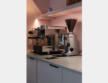 Eine moderne Kaffeezubereitungsstation mit einer großen Espressomaschine aus Edelstahl, daneben eine silberne Kaffeemühle mit transparentem Bohnenbehälter. Die Einrichtung umfasst eine rosa Wand und eine Arbeitsfläche mit Hängeregalen darüber.