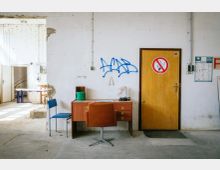 Ein leerer Raum in einem heruntergekommenen Gebäude mit einem weißen, teilweise beschädigten Putz an den Wänden. Im Vordergrund steht ein Holztisch mit einem braunen Stuhl und daneben ein blauer Stuhl; an der Wand befindet sich eine graue Tür mit einem "Rauchen verboten"-Schild darüber.