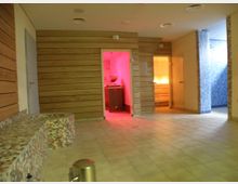 Ein moderner Wellnessbereich mit gefliesten Wänden und Böden. Im Vordergrund eine Sitzbank mit Mosaikfliesen, im Hintergrund Türen zu beleuchteten Räumen, darunter ein rot beleuchteter Raum und eine Duschkabine mit blauen Fliesen.