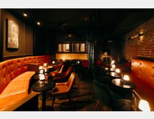 Dunkler Bar- oder Lounge-Innenraum mit orangefarbenen, gepolsterten Sitzbänken, runden schwarzen Tischen und kleinen Tischlampen. An der Seite ist eine freiliegende Backsteinwand, im Hintergrund eine Theke mit Spiegeln und gedämpfter Deckenbeleuchtung.