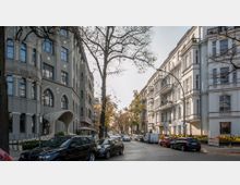 Straßenszene in einer städtischen Umgebung mit mehrstöckigen Gebäuden auf beiden Seiten. Links ein graues Gebäude mit spitzbogigen Fenstern und einem Vordach, rechts helle historische Fassaden mit Balkonen; am Straßenrand geparkte Autos und herbstlich gefärbte Bäume.