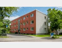 Mehrfamilienhaus mit drei Etagen, roten und weißen Fassaden sowie rechteckigen Fenstern. Vor dem Gebäude befindet sich eine gepflasterte Straße, ein Parkplatzschild sowie Grünflächen mit Bäumen.