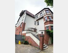 Mehrstöckiges Wohngebäude mit einer Kombination aus hell verputzten und roten Backsteinfassaden, einem Erker mit Fachwerk-Elementen und einem roten Schrägdach. Im Vordergrund führt eine Treppe mit Geländer zu einem Eingang, umgeben von Pflanzen und einem kleinen gepflasterten Hof.