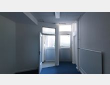 Ein schlichter Innenraum mit weißen Wänden, blauem Teppichboden und einer Tür aus Glas und Kunststoff, die ins Freie führt. Rechts an der Wand befindet sich ein Heizkörper, und der Raum wird durch eine Deckenleuchte beleuchtet.