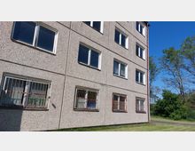 Mehrstöckiges Wohngebäude aus grauem Beton mit rechteckigen Fenstern. Einige Fenster im Erdgeschoss haben rostfarbene Gitter, im Hintergrund sind Bäume und ein blauer Himmel sichtbar.