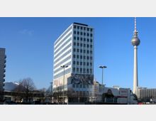 Ein modernes, rechteckiges Bürogebäude mit Glasfassade und Wandkunst im unteren Bereich, links umgeben von kahlen Bäumen. Im Hintergrund steht der Berliner Fernsehturm vor einem klaren blauen Himmel.