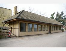 Flaches Gebäude mit Satteldach und gelblicher Fassade, das eine Reihe von rechteckigen Fenstern aufweist. Im Hintergrund stehen Bäume und ein großes, gelbes Gebäude.