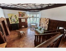 Großes Wohnzimmer mit Holzboden und dunkler Holzvertäfelung an den Wänden, dekoriert mit modernen Gemälden. Der Raum verfügt über eine Sitzecke mit Sofa, Sessel und Tisch, einen runden Esstisch mit Stühlen vor einem großen Erkerfenster, und eine hölzerne Treppe im Vordergrund.