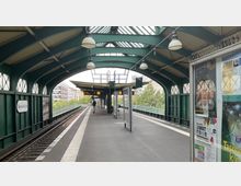 Überdachter Bahnsteig der U-Bahn-Station "Eberswalder Straße" mit grüner Stahlkonstruktion, einem Gleis und Sitzbereichen. Im Hintergrund Stadtansicht mit Bäumen und Gebäuden, rechts ein Schaukasten mit Plakaten.