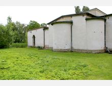 Außenansicht eines einfachen historischen Gebäudes mit cremefarbenen, verputzten Wänden und halbrunden Apsiden, flankiert von einer Wiese und umgeben von dichtem Grün. Das Gebäude weist eine kleine Rundbogentür und Ziegelsteinsockel auf.