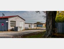 Industriegelände mit mehreren grauen einstöckigen Lagergebäuden, gepflasterter Straße und einem großen Baum im Vordergrund. Im Hintergrund sind Bäume und ein Zaun sichtbar, während ein Straßenschild „Betriebshof“ zeigt.