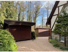 Gehweg aus Pflastersteinen, flankiert von einem Holzschuppen mit dunkelbrauner Fassade und einem Fachwerkhaus mit weißer Wand und braunen Holzbalken. Im Hintergrund stehen hohe, kahle Bäume, umgeben von grünen Sträuchern und Nadelbäumen.