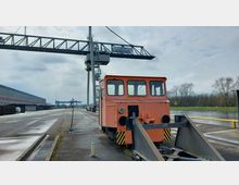 Kleiner orangefarbener Rangierzug auf einer Schienenstrecke am Stadthafen Brandenburg an der Havel, mit einem großen Stahlkran und Lagerhallen im Hintergrund. Im rechten Bildbereich fließt ein Fluss, umgeben von kahlen Bäumen.