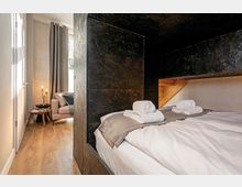 Modernes Schlafzimmer mit großem Bett, weißer Bettwäsche und Handtüchern, eingerahmt von einer dunklen, strukturierten Wandverkleidung und einem Holzelement am Kopfteil. Daneben ein kleiner Wohnbereich mit Sofa, Beistelltisch und Lampe sowie hohen Fenstern mit Vorhängen und hellem Holzboden.